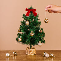 The Darling Effect Green Mini Artificial Christmas Tree, decorated with gold and white ornaments and a red bow topper, sits on a wooden surface. A hand adds a gold ornament; extra ornaments are nearby for festive decorating fun.