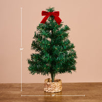 The Darling Effect Green Mini Artificial Christmas Tree, 16 inches tall with a red velvet bow and woven basket base, sits on a wooden table against a peach background—ideal for festive holiday decor.