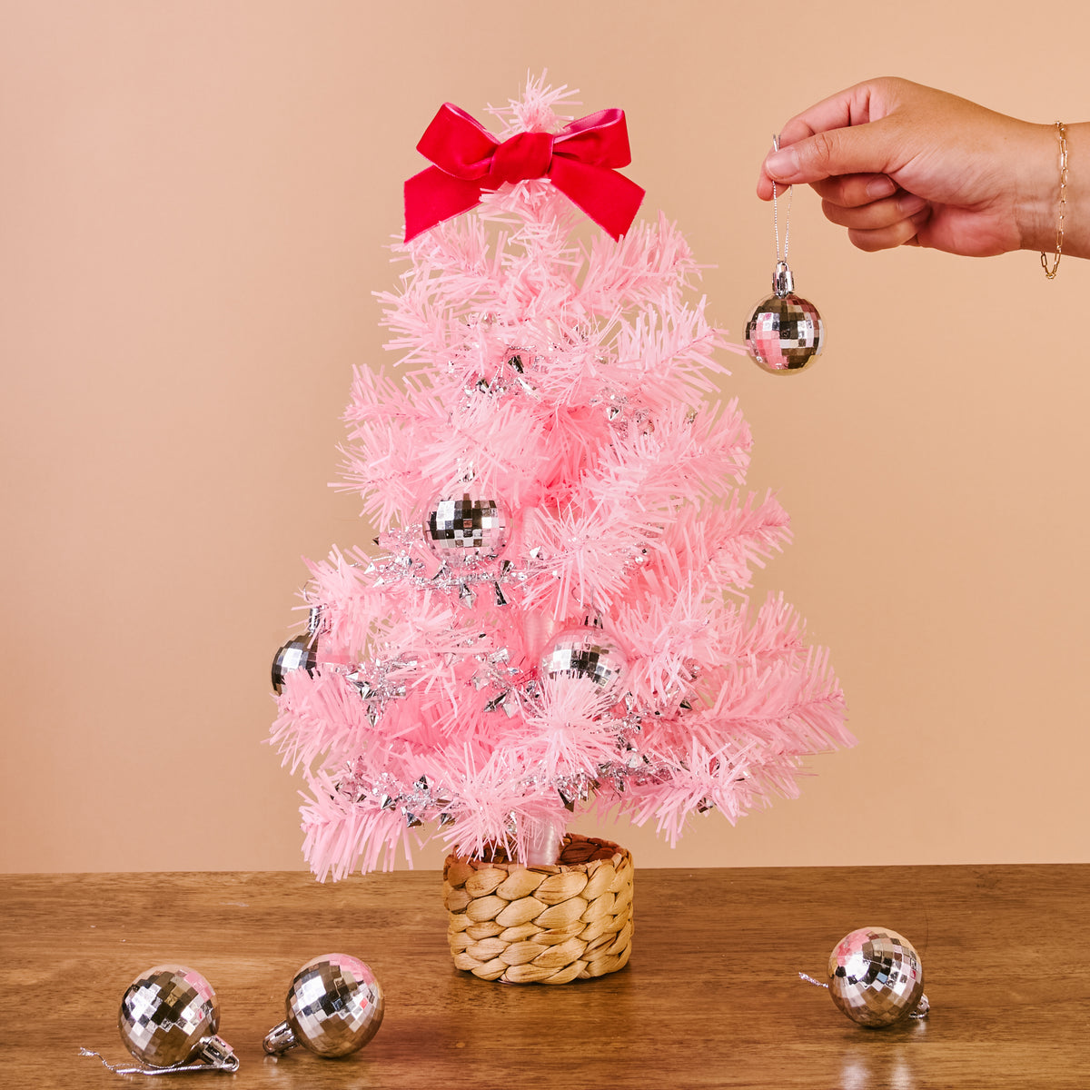 A hand hangs a silver disco ball ornament on a Pink Mini Artificial Christmas Tree by The Darling Effect that features a red bow topper.