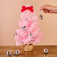 A hand hangs a silver disco ball ornament on a Pink Mini Artificial Christmas Tree by The Darling Effect that features a red bow topper.