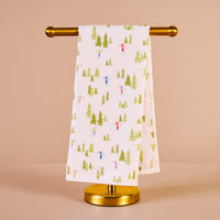 The Darling Effect Aprés Ski Waffle Tea Towel, featuring green trees and colorful skiers, is neatly draped over a gold towel stand against a beige background.