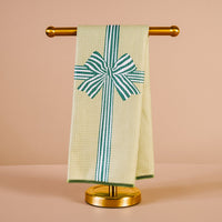 The Darling Effect Holiday Gift Bow Green Waffle Tea Towel, featuring a green striped bow and ribbon design, hangs on a gold towel stand against a light beige background.