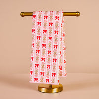The Darling Effect's Oh So Merry Waffle Tea Towel, featuring a red bow and floral pattern, hangs neatly on a gold stand against a beige background—an absorbent and charming kitchen accessory.