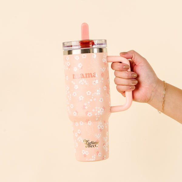 A hand holds the-darling-effect's Mama Peach 40 oz Lifestyle Flip Straw Tumbler, featuring a pink floral print, “mama” script, pink lid and handle, flip straw, and logo on the bottom—stylish and leak-resistant.