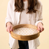 A person in a white shirt holds the-darling-effect Handpicked For You Large Round Wood Enamel Tray, featuring a pink and yellow geometric floral pattern, against a plain beige background. The person’s head is not visible.