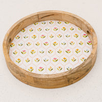 The-darling-effect’s Handpicked For You Large Round Wood Enamel Tray features short sides, cut-out handles, and a white base with a hexagonal colorful floral pattern, displayed on a white speckled surface.