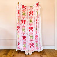 The Darling Effect's Alpha Omicron Pi Plush Sherpa Blanket in light pink features pink flowers, green leaves, red bows, and "alpha omicron pi" repeated vertically—perfect for a college dorm throw.