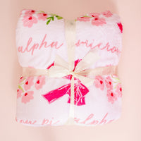 The Darling Effect’s Alpha Omicron Pi Plush Sherpa Blanket, featuring pink floral patterns and cursive pink text, is folded with a cream ribbon bow—perfect as a soft college dorm throw against a pale pink background.