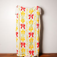 A person stands behind The Darling Effect's Chi Omega Plush Sherpa Blanket, featuring red bows, yellow flowers, and cursive script. Only their hands and feet show as the cozy blanket contrasts with a white wall and wooden floor.