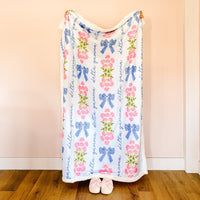 Wrapped in The Darling Effect’s Delta Gamma Plush Sherpa Blanket with blue bows, pink flowers, and cursive text, only their pink slippers are visible on a wooden floor against a pale pink wall.