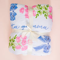 A soft, white Delta Gamma Plush Sherpa Blanket by The Darling Effect, decorated with pink flowers, green leaves, and blue bows, is neatly folded and tied with a cream ribbon. “Delta Gamma” text is partially visible on this cozy dorm throw.