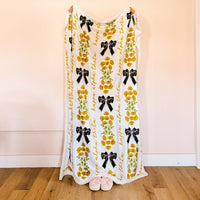 A person stands on a wooden floor, mostly hidden behind The Darling Effect's Kappa Alpha Theta Plush Sherpa Blanket featuring yellow flowers and black bows. Only their hands and pink slippers peek out from this cozy sorority blanket.