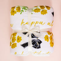 The Darling Effect's Kappa Alpha Theta Plush Sherpa Blanket—white with yellow flowers and cursive text—is neatly folded, tied with a cream ribbon, and displayed on a pale pink background, making it a cozy college dorm throw.