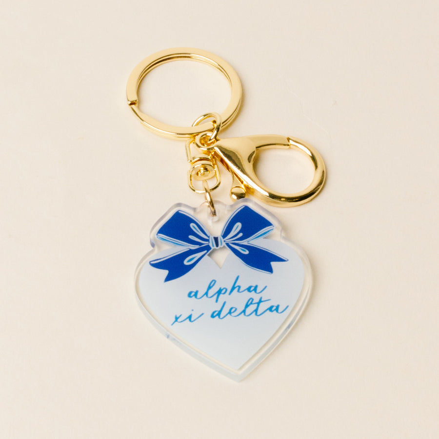 The Darling Effect Alpha Xi Delta Sweetheart Bow Acrylic Keychain features a heart shape, blue script, pastel bow, and gold keyring with clasp—perfect as a sisterhood gift.