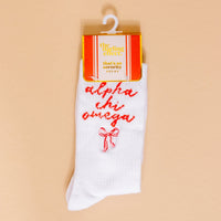 The Darling Effect Alpha Chi Omega Embroidered Crew Socks feature "alpha chi omega" in red script with a small red bow. Displayed on beige and packaged as That's So Sorority Socks—perfect for unique sorority gifts.