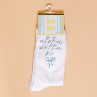 Show off your sorority spirit with The Darling Effect's Alpha Delta Pi Embroidered Crew Socks—white socks featuring blue script, a cute blue bow, and a yellow "the darling effect thats so sorority socks" label, displayed on a beige background.
