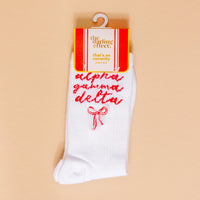 Show your sorority spirit with The Darling Effect Alpha Gamma Delta Embroidered Crew Socks—white crew socks featuring red script and a red bow, packaged on a peach background.