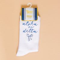 Show your sorority spirit with Alpha Xi Delta Embroidered Crew Socks from The Darling Effect—white crew socks featuring “alpha xi delta,” a blue bow, and cute packaging, perfect for any member.