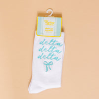 The Darling Effect’s Delta Delta Delta Embroidered Crew Socks feature teal cursive lettering and a teal bow—perfect for sorority gifts. Displayed on a peach background with retail tag.