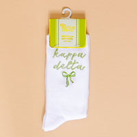 Kappa Delta Embroidered Crew Socks by The Darling Effect: white crew socks with green "kappa delta" text and a green bow, displayed on beige with yellow “The Darling Effect” packaging—perfect for thoughtful sorority gifts.