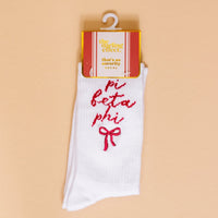 A pair of Pi Beta Phi Embroidered Crew Socks by The Darling Effect, featuring white fabric, pink bow, and sorority name on a peach background—perfect for showing off your Pi Beta Phi spirit.