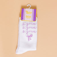 The Darling Effect’s Sigma Sigma Sigma Embroidered Crew Socks feature “sigma” in purple cursive three times with a small purple bow—displayed on peach background and packaging labeled “that’s so sorority socks”—perfect for sorority gifting.