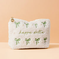 The Kappa Delta Bows & Pearls Teddy Pouch by The Darling Effect features green embroidered bows, pearl accents, and “kappa delta” in green on soft white sherpa—an ideal sorority gift, pictured on a light peach background.