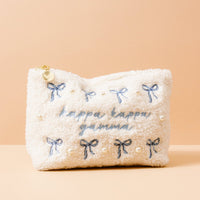 The Kappa Kappa Gamma Bows & Pearls Teddy Pouch by The Darling Effect is a white sherpa pouch featuring blue embroidered bows, pearls, and "kappa kappa gamma" in blue—perfect as a sorority gift. Set against a light peach background.