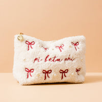 The Darling Effect's Pi Beta Phi Bows & Pearls Teddy Pouch is a white sherpa pouch with red embroidered bows, pearl accents, and "pi beta phi" stitched in red on the front—a sweet sorority gift against a beige backdrop.