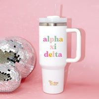 The Darling Effect Alpha Xi Delta 40 oz Take Me Everywhere White Tumbler, featuring colorful Alpha Xi Delta text and a handle with straw, sits beside a disco ball on a pink background. "The Darling Effect" is written at the bottom.