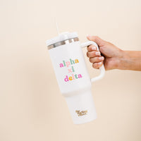 A hand holds The Darling Effect's Alpha Xi Delta 40 oz Take Me Everywhere White Tumbler, featuring a large handle, straw, colorful Alpha Xi Delta letters, and "the lifting effect" in gold near the bottom.