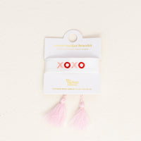 The XOXO Woven Bracelet by the-darling-effect features white woven fabric, pink and red embroidered "XOXO," two light pink tassels, and comes on a white card with gold text.