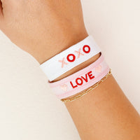 A hand wears the-darling-effect’s XOXO Woven Bracelet—white with red “XOXO”—paired with a pink LOVE bracelet and a gold chain, perfect for stacking colorful, trendy styles with embroidery accents.