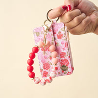 A hand with red nail polish holds The Darling Effect’s Rose Garden Keychain Wallet, a pink floral slim wallet card holder with zipper, gold keyring, and a beaded keychain in red, pink, and white—ideal for stylish organization.