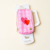 A stylish pink checkered fanny pack, the Charming Gingham Tumbler Fanny Pack by The Darling Effect, features two heart designs, a gold zipper, and a round gold emblem for a chic look, displayed on a pale surface.
