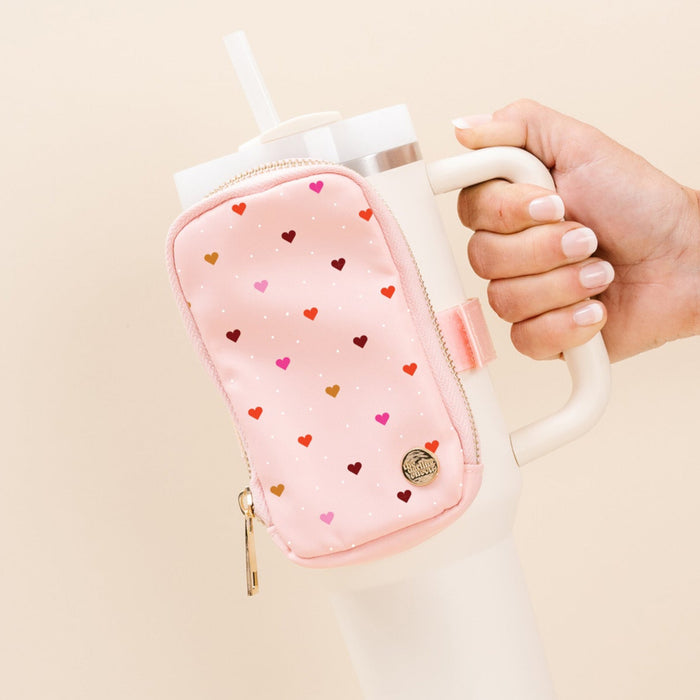 Hand holding a white mug with a pink heart-patterned sleeve against a beige background