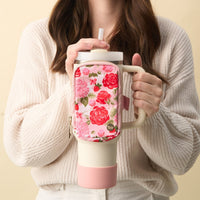 A woman in a cream sweater holds a large cup with a handle and straw, featuring The Darling Effect's Rose Garden Tumbler Fanny Pack—a water-resistant pink and floral zippered pouch attached to the front.