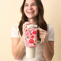 A smiling woman with long brown hair holds a large cream tumbler with a straw, featuring The Darling Effect's Rose Garden Tumbler Fanny Pack—a stylish pink floral accessory—attached to the handle. She wears a short-sleeved, ribbed white top.