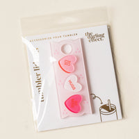 The Darling Effect Candy Hearts Tumbler Lid Tag features three pastel heart charms with "XOXO," "HUGS," and "BE MINE" messages. Perfect for personalizing your Stanley Quencher 2.0, with branded packaging included.