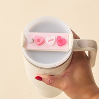 A hand with red nail polish holds a white mug of milky drink, topped with The Darling Effect's Candy Hearts Tumbler Lid Tag featuring three candy hearts labeled XOXO, HUGS, and BE MINE.