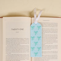 A delightful gift for book lovers, the-darling-effect’s Bows Blue Acrylic Bookmark features a green bow, white dot accents, and a white ribbon, shown atop an open book turned to chapter twenty-one on a light surface.