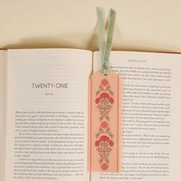 An open book displays chapter twenty-one with The Darling Effect’s Everbloom Dusty Rose Acrylic Bookmark—featuring red flowers, two ribbon ties, and a velvet tassel—placed on the right page; a perfect gift for book lovers.