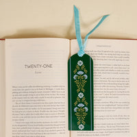 An open book displays The Darling Effect's Everbloom Forest Green Acrylic Bookmark, adorned with blue flowers and a light blue velvet ribbon tassel, placed vertically—an ideal gift for book lovers.