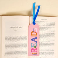 An open book displays chapter twenty-one with The Darling Effect's Read Acrylic Bookmark—featuring colorful "READ" letters and a velvet ribbon tassel—resting on the right page, making a charming accessory for book lovers.