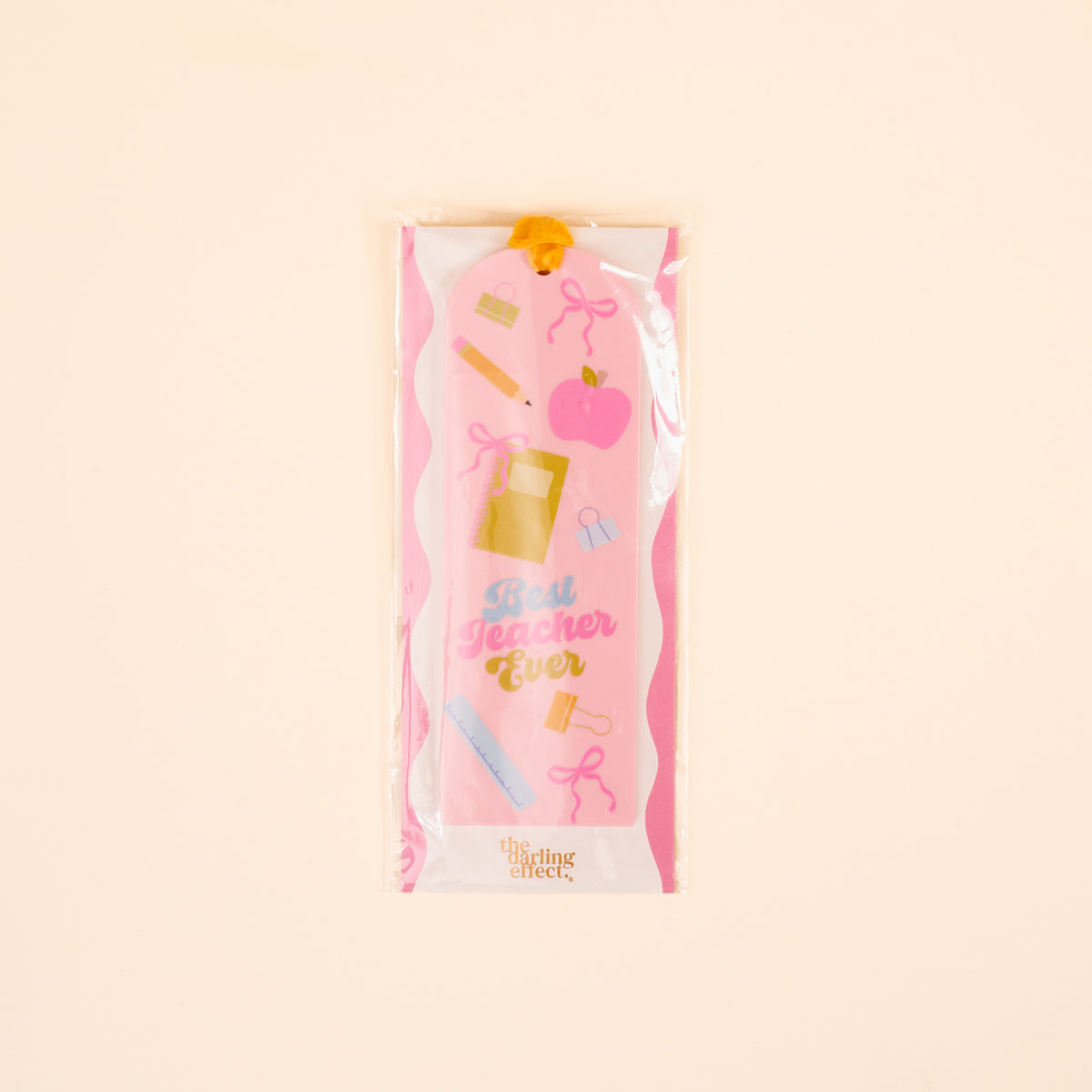 The Darling Effect's Best Teacher Ever Acrylic Bookmark features pink and white colors, school-themed illustrations, and “Best Teacher Ever” text. It comes in clear plastic packaging with a yellow ribbon, making it a perfect gift for book lovers.