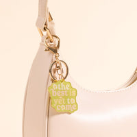 A cream-colored handbag with a gold clasp is styled with the-darling-effect’s The Best Is Yet to Come Acrylic Keychain, featuring yellow and green lettering for a cheerful, chic accessory.