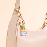 A close-up of a cream-colored handbag featuring The Darling Effect's Book Nook Acrylic Keychain—an accessory shaped like a stack of colorful books with a small green plant, accented by a gold zipper.