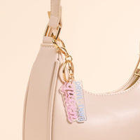 The Drive Thru Queen Acrylic Keychain by the-darling-effect, in pastel pink and blue, hangs from a beige shoulder bag—an adorable accessory for fans of cute keychains.