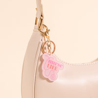 A close-up of a white handbag with the-darling-effect's Nurse Life Acrylic Keychain attached, featuring a pink and white medical badge charm and bold "nurse LIFE" lettering.