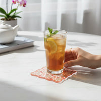 A hand holds a glass of iced tea with lemon and mint on The Darling Effect's Everbloom Dusty Rose Table Talk Acrylic Coaster, atop a white table. Behind it: a teapot, books, and an orchid complete the elegant scene.